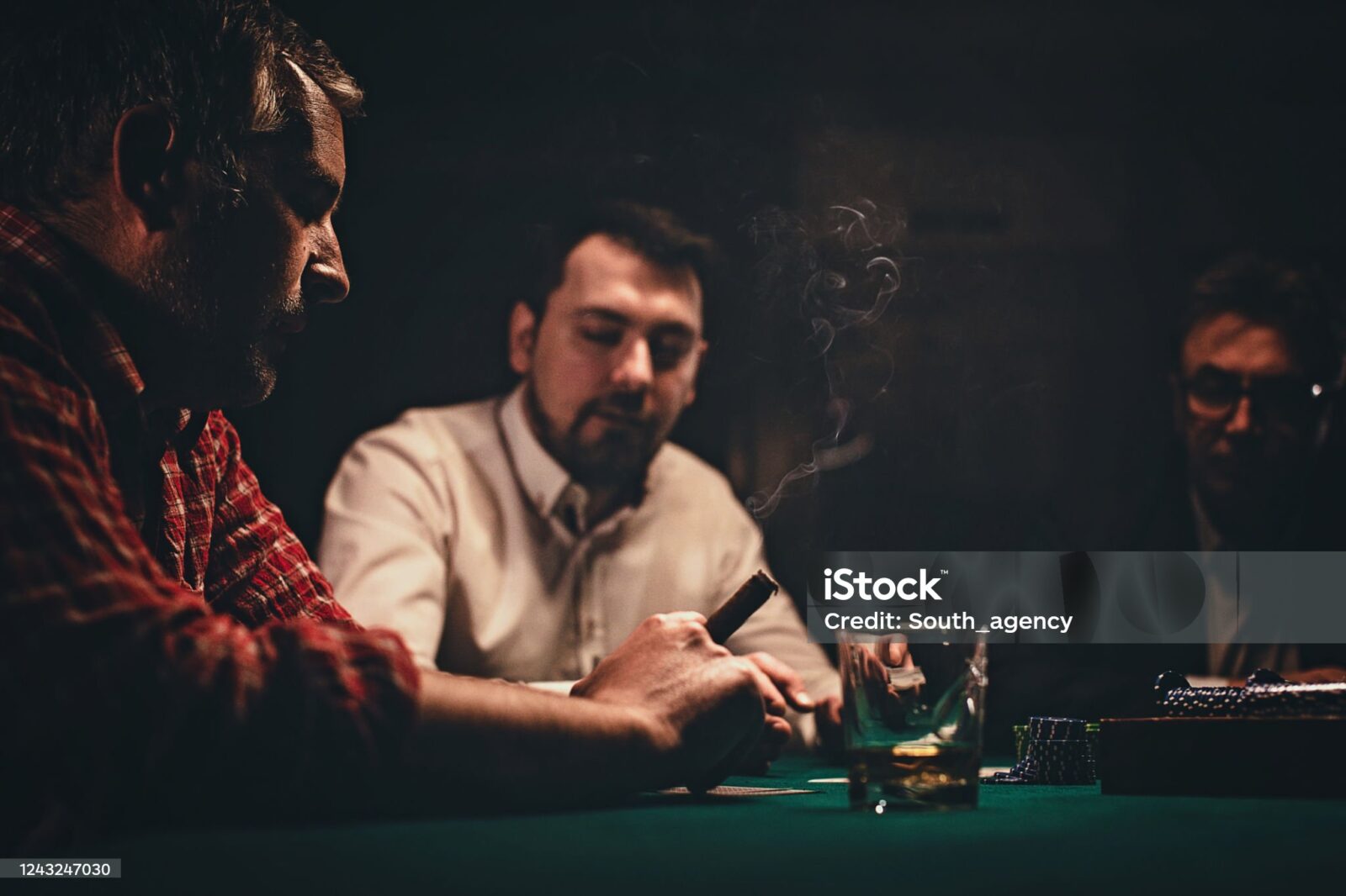 Three people playing cards at a dimly lit table, cigar smoke rising