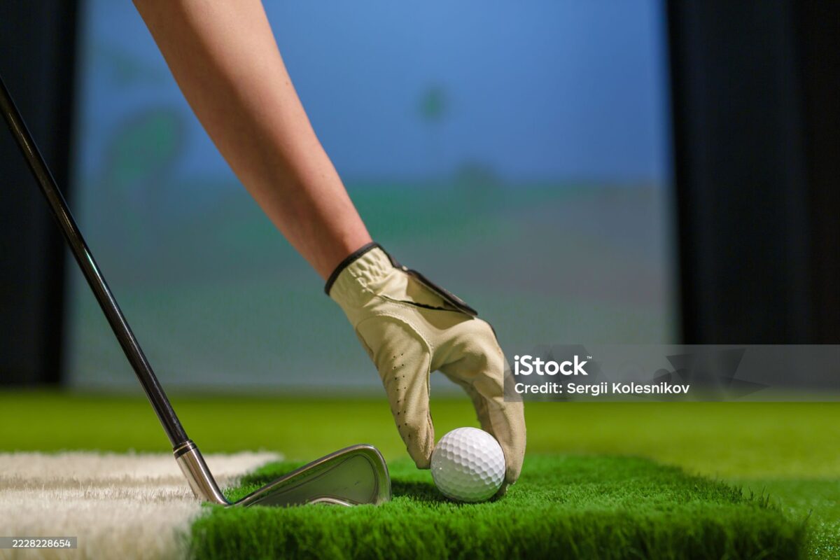 A gloved hand placing a golf ball beside an iron on simulator turf